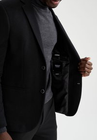 Musta bleiseri, jossa on teksturoitu pinta, kaksi nappia ja slim fit -leikkaus. Sisäpuolella mustasta kankaasta vuoritettu, jossa etiketti ja huomaamattomat taskut.