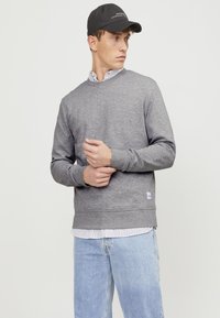 Gray sweatshirt with long sleeves and ribbed cuffs, worn over a striped collared shirt, paired with light blue jeans and a black cap.
