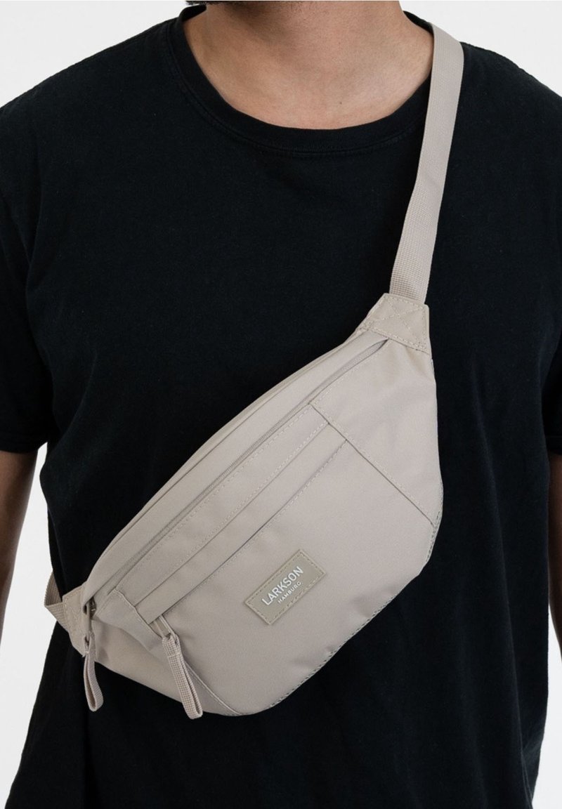 Personne portant un sac bandoulière beige avec deux fermetures éclair et une étiquette Larkson sur une chemise noire, visible du cou à la taille.