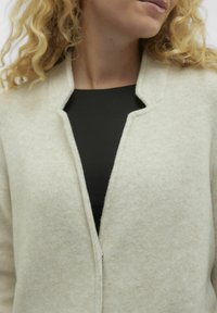 Giacca in pile di un beige chiaro con una finitura testurizzata, presenta un design aperto davanti e un colletto arrotondato. Indossata sopra una maglietta nera.