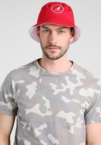 Cappello bucket rosso Kangol con una visiera interna bianca, caratterizzato da una toppa circolare con logo. Il modello indossa una t-shirt grigia a maniche corte con motivo camouflage.