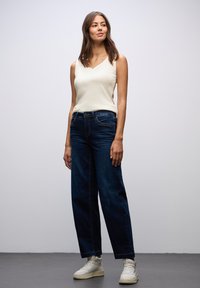 Dark blue wide-leg jeans paired with a light beige sleeveless top. The model stands on a neutral background, wearing white sneakers.