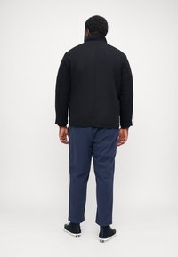 Black quilted jacket with a high collar, worn with navy trousers and black sneakers. Features a sleek, modern silhouette.