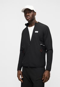 Άνδρας που φοράει μαύρο μπουφάν με φερμουάρ και λογότυπο EA7, λευκό πουκάμισο, μαύρο παντελόνι και λευκό καπέλο του μπέιζμπολ, στέκεται μπροστά σε ένα απλό γκρι φόντο.