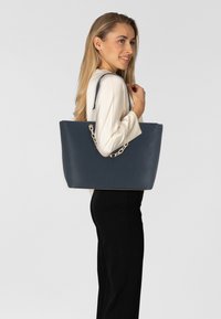 Námorně modrá, texturovaná kožená taška přes rameno s dvojitými ramenními popruhy a řetízkovým detailem, obdélníkový tvar a otevřený design.