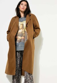 Brown wollen jas met een ontspannen pasvorm, knopen aan de voorkant. Daaronder een graphic sweater met de Mona Lisa, gecombineerd met een patroonbroek.
