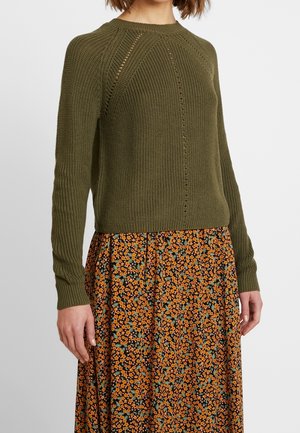Woman wearing an olive-green knit sweater with long sleeves paired with a black skirt featuring an orange floral pattern.