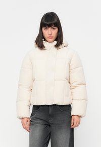 Kvinne med rett mørkt hår iført en kremfarget dunjakke med hette og mørkegrå høytlivsjeans som står mot en hvit bakgrunn.