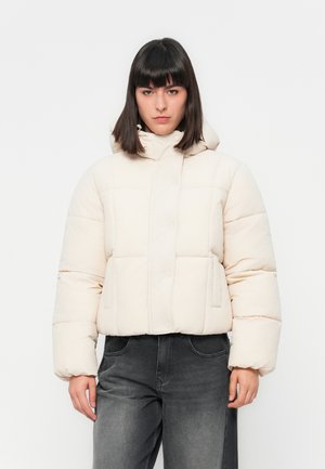 Femme avec des cheveux raides et foncés portant une veste en duvet crème avec capuche et un jean taille haute gris foncé, se tenant contre un fond blanc.