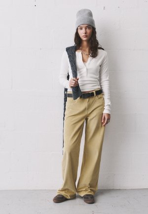 Hvid langærmet henley-top, beige corduroy bukser med vidde og bælte, grå strikket hue og brune slip-on sko. Holder en mørk vest.