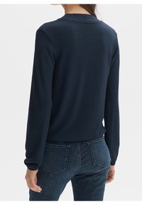 Námořnická modrá halenka s dlouhými rukávy z jemného materiálu, se zaobleným výstřihem a mírně vypasovaným střihem, kombinovaná s tmavými džíny a zadními kapsami.