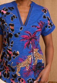 Kék, rövid ujjú, V nyakú ing, rózsaszín, fekete és bézs leopárd és pálmafa mintázattal. Hímzett díszítések az ujjak mentén.