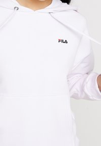Felpa bianca realizzata in tessuto morbido con tasca frontale, decorata con un piccolo logo FILA nero con accenti rossi e blu vicino al collo.