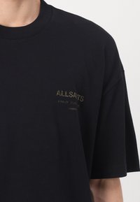 Fekete rövid ujjú póló, bal mellrészen kis arany "ALLSAINTS" logóval és japán szöveggel, fehér háttér előtt egy személy által viselve.