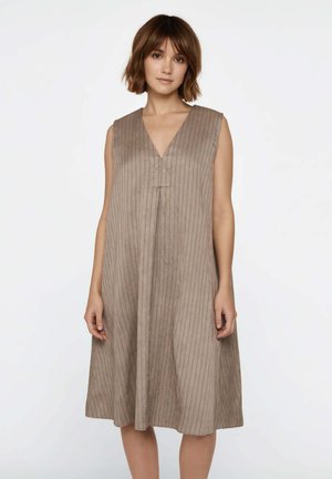 Donna con capelli castani corti che indossa un vestito beige senza maniche con righe verticali e scollo a V, in piedi su uno sfondo bianco uniforme.