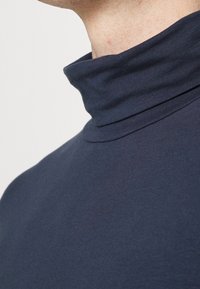 Laivastonsininen korkeakauluksinen paita pehmeästä kankaasta, jossa on korkea kaulus ja sileä pinta, kuvattu sivulta.