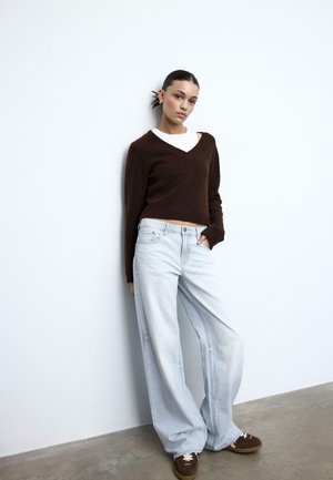 Mladá žena opierajúca sa o stenu, oblečená v hnedom svetri s výstrihom do V, bielom topu, svetlých širokých džínsoch a hnedých teniskách, s vlasmi zviazanými dozadu.