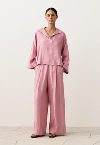 Nő áll laza illeszkedésű, rózsaszín len felsőt viselve, széles gallérral és hozzáillő, bő szárú nadrágot, kezét a zsebében, fekete szandált visel csempézett padlón.