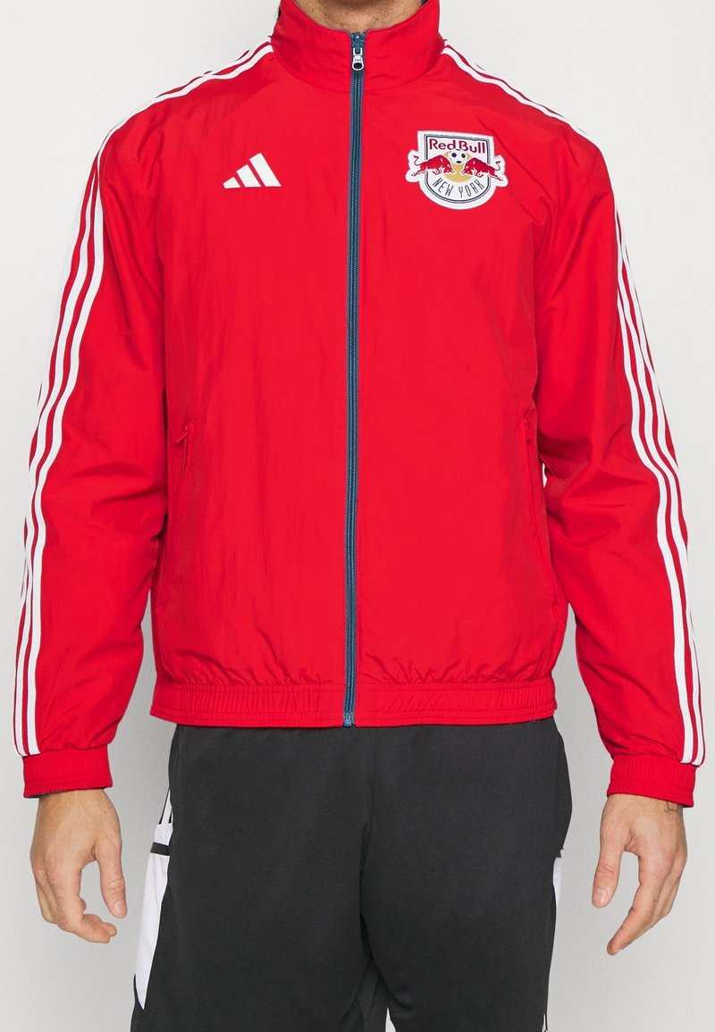Rote Jacke aus leichtem Stoff, mit Reißverschluss vorne, hohem Kragen und weißen Streifen an den Ärmeln. Logo auf der linken Brust.