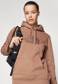 Felpa con cappuccio marrone, caratterizzata da un grande logo sulla parte frontale, tasca a marsupio e una finitura testurizzata. Borsa nera con tracolla regolabile in mano.