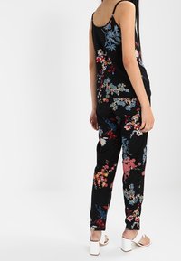 Zwarte bloemige jumpsuit met kleurrijke patronen; heeft een mouwloze top en taps toelopende broeken. Verfraaid met fijne bloemenborduursels.