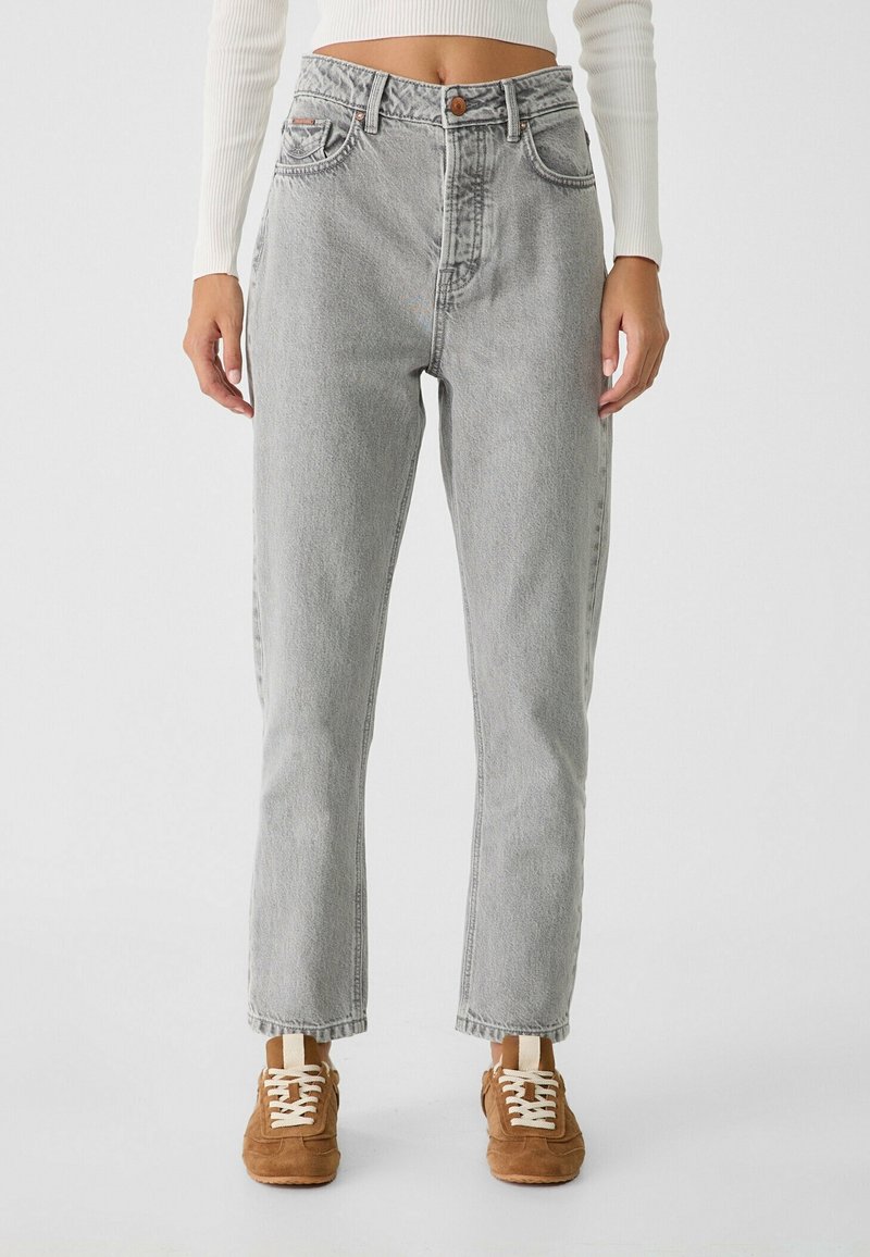 Persona que lleva unos pantalones rectos de talle alto en gris claro, una camiseta de manga larga blanca acanalada y zapatillas de gamuza marrón con cordones blancos.