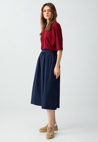 Jeune femme portant un haut rouge texturé, une jupe midi bleu marine et des baskets compensées beige, debout devant un fond clair uni.