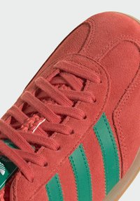 Červené semišové tenisky s textúrovaným povrchom, zelenými koženými prúžkami, zodpovedajúcimi šnúrkami a svetlohnedou gumovou podrážkou.