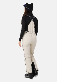 Beige skidragter med justerbare stropper, sorte sideakcenter og lynlåsdetailler, parret med en sort langærmet bluse og en sort hue.