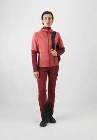 Naiselg seisab seljas kahekohaline punane lukuga jakki, sobivate pükstega, mille mustad alumised osad on mustad, mustad kingad ja must seljakoti rihm üle õla.