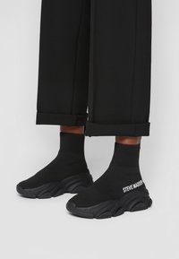Baskets de style chaussette noires avec une semelle épaisse et texturée. Caractéristiques d'une cheville côtelée et d'un logo "STEVE MADDEN" blanc sur le côté.