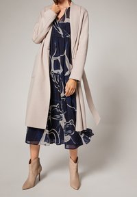 Beiger Wollmantel mit langen Ärmeln, offenem Vorderteil und Seitentaschen, getragen über einem marineblauen Kleid mit abstrakten, hellfarbenen Tiermustern. Beigefarbene Knöchelstiefel.