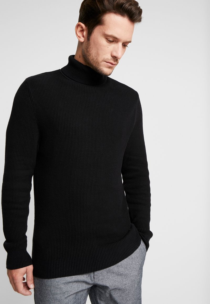 Suéter negro de punto acanalado con cuello alto y mangas largas, que presenta un diseño ajustado y una ligera variación en el dobladillo. Textura suave, sin patrones visibles.