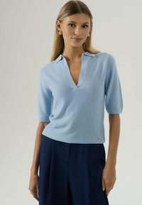 Žena v svetlomodrom krátkom rukáve pletenom topu s výstrihom do V a námorníckych širokých nohaviciach, stojí pred jednoduchým pozadím.