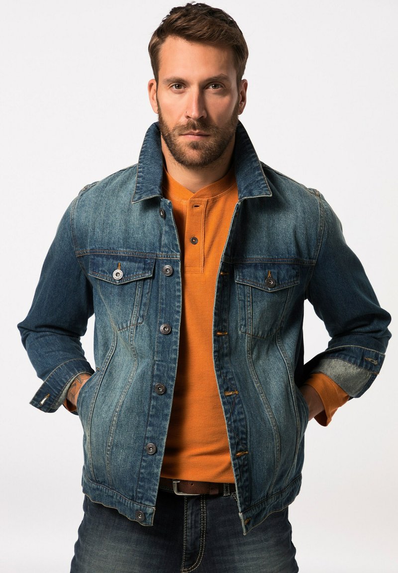Chaqueta de mezclilla con frente abotonado, dos bolsillos en el pecho y color azul desgastado. Usada sobre una camisa de manga larga naranja; diseño casual con cuello estructurado.