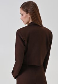 Bruine cropped blazer met gestructureerde schouders, een gladde textuur en verborgen sluitingen, gezien vanaf de achterkant. Lang haar met een hangende oorbel.