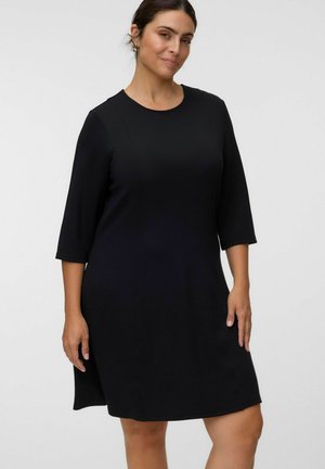 Femme portant une robe noire mi-longue avec des manches trois-quarts, debout devant un fond uni clair.