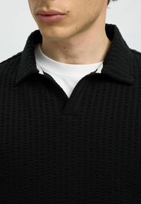 Juodas tekstūruotas megztinis su storu, raižytu raštu ir įpjautu apykakle, uždėtas ant baltos apvalios iškirptės marškinėlių.