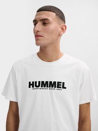 Joven con el pelo corto que lleva una camiseta blanca con la inscripción "HUMMEL SPORTSWEAR SINCE 1923" impresa en negro en la parte delantera.
