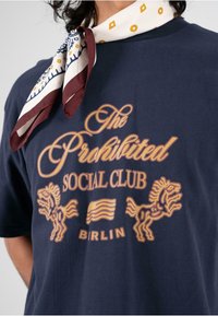 Jūros laivyno medvilnės marškinėliai su aukso spalvos tekstu "Draudžiamas socialinis klubas BIRLIN" ir liūto grafika, papildyti spausdinta šalikų aplink kaklą.