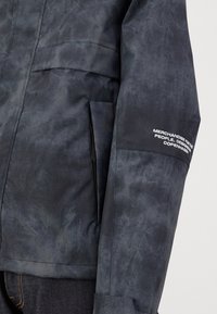 Waterafstotende zwarte jas met een gestructureerde afwerking, voorzien van zakken en witte bedrukte tekst op de mouw. Minimalistische designelementen.