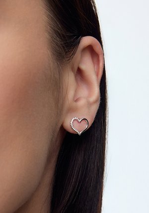 Srebrne kolczyki w kształcie serca z przezroczystym bordurem, otwartym centrum i teksturowanymi detalami, noszone na uchu modelki.