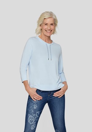 Hellblauer Sweatshirt mit rundem Halsausschnitt und Zugbandakzenten, kombiniert mit dunkelblauen Jeans, die ein florales Muster am linken Bein aufweisen.