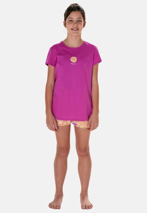 Junges Mädchen steht barfuß da, trägt ein lila T-Shirt mit einem Orangen-Scheiben-Motiv und orangemgemusterte Shorts vor weißem Hintergrund.