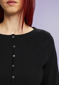 Musta pitkähihainen paita, jossa on pyöreä pääntie ja napitus, jossa on viisi näkyvää nappia. Suora, joustava kangas ilman kuvioita.