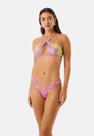Donna che indossa un bikini floreale rosa e giallo con top a incrocio halter e slip bikini a vita alta, in piedi contro uno sfondo chiaro e uniforme.