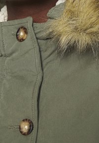 Olivgrön jacka med fuskpäls vid huven, stora knappstängningar och quiltad textur. Materialet verkar hållbart med robusta sömmar.