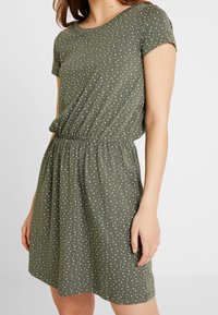 Robe verte à pois en tissu doux, avec manches courtes, taille élastique et poches latérales. Motif à pois blancs.