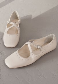 Krém színű bőrből készült balerinacipők keresztpántokkal, négyzet alakú orral, és díszítő fém szegecsekkel a széleken. Lapos talpúak, letisztult, minimalista dizájnnal.