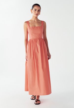 Coral, sleeveless maxi dress with a square neckline and gathered waist. Made of smooth fabric, it features a flowing skirt and black sandals.